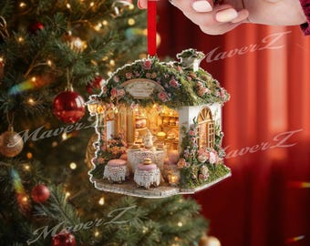 Puede incluir: Adorno navideño con forma de panadería en miniatura, adornado con rosas rosas y vegetación. El adorno presenta un interior detallado con pasteles y mesas en miniatura. Se suspende con una cinta roja sobre un fondo de árbol de Navidad.