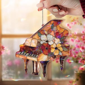 May include: A stained glass grand piano ornament with a floral design. The piano is a warm amber color with a keyboard and legs. The flowers are white, red, and yellow. The ornament is held by a chain.