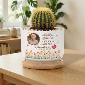May include: A white ceramic planter with a wooden base containing a green cactus. The planter features a circular photo of an older person, yellow butterflies, a red heart, and the text: Because someone we love is in heaven there's a little bit of Camellia in our home, above a colourful floral border.