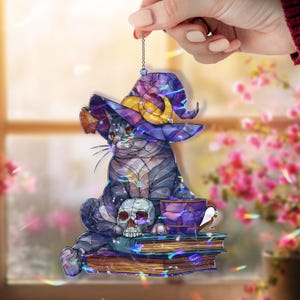 Puede incluir: Figura decorativa de gato con sombrero de bruja, sentado sobre libros con una calavera y una taza de té. El gato es gris y azul con un sombrero morado y detalles dorados. La figura se sujeta con una cadena.