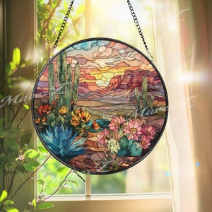 May include: A round stained glass window featuring a desert landscape with cacti, flowers, and a sunset. The artwork uses shades of blue, orange, pink, and green, with a black metal frame and hanging chain.