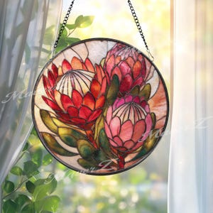 May include: A circular stained glass suncatcher featuring three protea flowers in shades of red, pink, and orange, surrounded by green leaves. The piece is framed in a dark metal border and hangs from a black metal chain against a bright window.