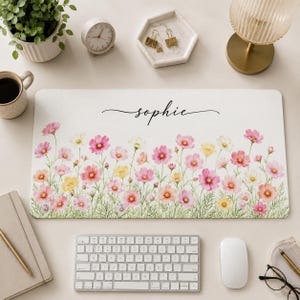 May include: A rectangular desk mat featuring a white background with a watercolor-style floral print of pink, yellow, and white cosmos flowers along the bottom edge. The name "sophie" is printed in a black cursive script in the upper center.