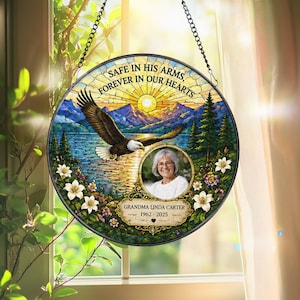 May include: A circular stained glass-style hanging ornament featuring a bald eagle flying over a lake with mountains and a sunburst. It includes a central circular photo frame, white lilies, and the text: SAFE IN HIS ARMS, FOREVER IN OUR HEARTS, GRANDMA LINDA CARTER, 1962-2025.