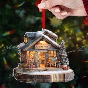 Pode incluir: Enfeite de Natal em miniatura em forma de loja rústica com o texto "ATUID STORLO" acima da entrada. O enfeite apresenta uma pequena árvore de Natal, um telhado coberto de neve e uma fita vermelha para pendurar. A loja está sobre uma base de madeira.
