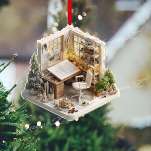 May include: A miniature architectural office ornament with a red ribbon for hanging. The ornament features a drafting table, chair, bookshelves, and small Christmas trees. The interior is lit with small lights, creating a cozy scene.