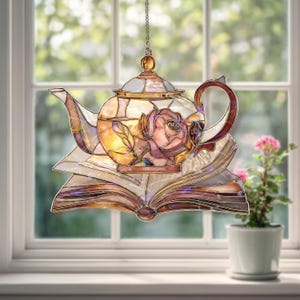 Puede incluir: Tetera de vidriera con diseño de rosa, colgada de una cadena. La tetera descansa sobre un libro abierto, con un arreglo floral en el fondo. El vidrio presenta tonos dorados, rosas y marrones.