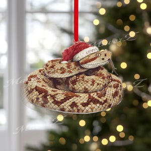 Puede incluir: Adorno navideño de serpiente enroscada con un gorro de Papá Noel rojo que dice "COVES". La serpiente tiene un patrón marrón y beige. El adorno cuelga de una cinta roja. El fondo está borroso con luces de árbol de Navidad.