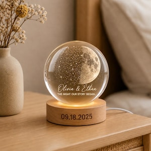 May include: A clear glass sphere lamp featuring an internal laser-etched moon and star design, engraved with the text Olivia & Ethan THE NIGHT OUR STORY BEGAN. The sphere rests on a round wooden base engraved with the date 09.18.2025.