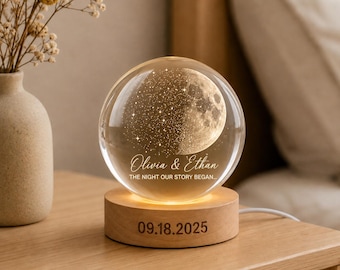 May include: A clear glass sphere lamp featuring an internal laser-etched moon and star design, engraved with the text Olivia & Ethan THE NIGHT OUR STORY BEGAN. The sphere rests on a round wooden base engraved with the date 09.18.2025.