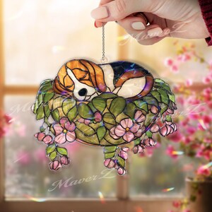 May include: A stained glass suncatcher depicting a sleeping beagle puppy in a bed of green leaves and pink flowers. The puppy has brown, white, and black markings. The suncatcher is suspended by a chain.