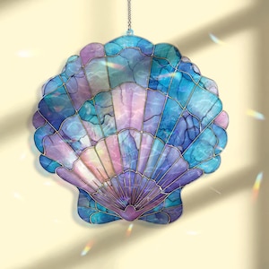 以下が含まれることがあります： 青、紫、ピンクの色合いのステンドグラスの貝殻サンキャッチャー。貝殻は金色の金属で縁取られ、チェーンから吊り下げられています。デザインは扇のような模様が特徴です。