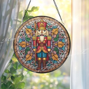 May include: Round stained glass window art featuring a nutcracker soldier in red, blue, and gold. The artwork is framed in a black metal ring and hangs from a chain. The background is a colorful mosaic with holly leaves.