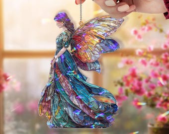 Op de afbeelding: Een kleurrijk feeënornament met iriserende vleugels en een vloeiende blauwe jurk. De fee is afgebeeld in een glas-in-loodstijl, met een ketting om op te hangen. De achtergrond is wazig, met roze bloemen en een raam.
