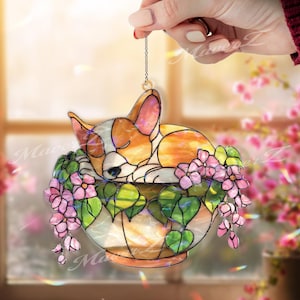 May include: Stained glass ornament featuring a sleeping corgi puppy in a flower pot. The corgi is orange, white, and brown. The pot is filled with pink flowers and green leaves. The ornament is hanging from a chain.