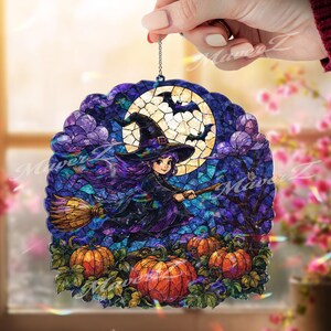 May include: A circular stained glass-style ornament featuring a witch with purple hair flying on a broomstick against a large yellow moon. The scene includes silhouettes of bats, purple clouds, and orange pumpkins on a dark blue background.