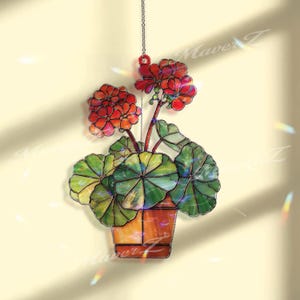 May include: Stained glass hanging art piece depicting a potted geranium plant. The piece features red flowers, green leaves, and a brown pot. The artwork is suspended by a chain and is designed to catch the light.