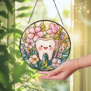 May include: A circular stained glass suncatcher featuring a central smiling tooth character surrounded by pink flowers, green leaves, and a pink toothbrush. The piece is framed in a dark metal border and hangs from a black metal chain.