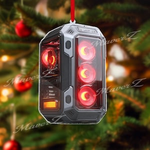 May include: A computer case Christmas ornament with a red ribbon for hanging. The ornament is grey and black with red glowing lights inside. The ornament is hanging in front of a Christmas tree.