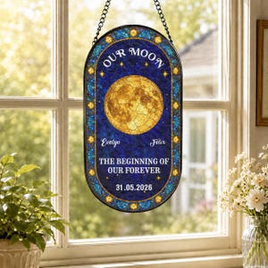 May include: An oval stained glass suncatcher featuring a golden moon against a deep blue background. The text reads OUR MOON, Evelyn, Felix, THE BEGINNING OF OUR FOREVER, and 31.05.2026. It has a decorative blue and gold border and a black chain.