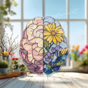 Può includere: Un acchiappasole in vetro colorato a forma di cervello umano. Il lato sinistro presenta toni tenui di rosa e viola con un motivo strutturato. Il lato destro mostra una margherita gialla brillante e fiori blu con foglie verdi.