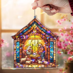 May include: A stained-glass style Christmas ornament depicting the Nativity scene. The ornament features a stable with Mary, Joseph, baby Jesus, and sheep. The ornament is held by a hand and has a chain for hanging.