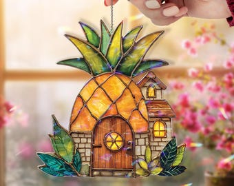May include: A stained glass pineapple ornament with a house design. The pineapple is yellow and orange with green leaves. The house has a wooden door and brick walls. The ornament is held by a hand.