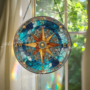 May include: A round stained glass compass with a gold and teal color scheme. The compass rose is centered with a gold star design. The background is a mosaic of teal and blue glass. The compass is hanging from a black chain.