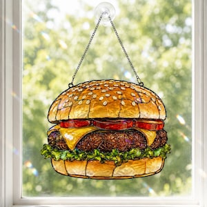 Op de afbeelding: Een glas-in-lood zonnevanger in de vorm van een cheeseburger, met een goudbruin broodje met witte sesamzaadjes, rode tomatenplakken, gele kaas, een donkerbruine burger en groene sla, opgehangen aan een metalen ketting met een zuignap.
