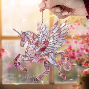 Może przedstawiać: Ozdoba w kształcie jednorożca ze szkła witrażowego w odcieniach różu, fioletu i bieli, ze srebrnym łańcuszkiem do zawieszenia. Mityczne stworzenie ma rozłożone skrzydła i spiralny róg. Ozdoba jest trzymana przez dłoń przed oknem z różowymi kwiatami.