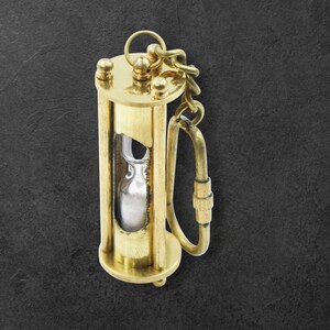 Può includere: Un piccolo portachiavi a clessidra in ottone con catena e chiusura. La clessidra ha un centro in vetro trasparente con sabbia color argento. L'involucro in ottone ha una forma cilindrica con dettagli decorativi.