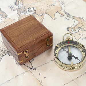 Może przedstawiać: Mosiężny kompas z białą tarczą i czarną igłą spoczywa na szczegółowej mapie świata. Obok kompasu znajduje się drewniane pudełko z mosiężnymi detalami. Kompas głosi "THE JOURNEY BEGINS WITHIN".