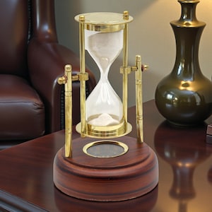 May include: An hourglass with a brass frame and a wooden base. The glass bulb contains white sand. The hourglass is approximately 7 inches tall. The base is a dark brown color. The hourglass is a decorative item.