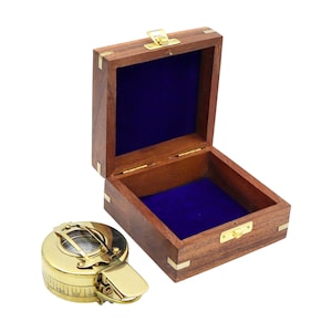 May include: A round brass compass with a hinged lid and a side latch, positioned next to an open square wooden box. The box features brass corner accents, a brass latch, and a deep blue velvet lining on the interior base and lid interior.