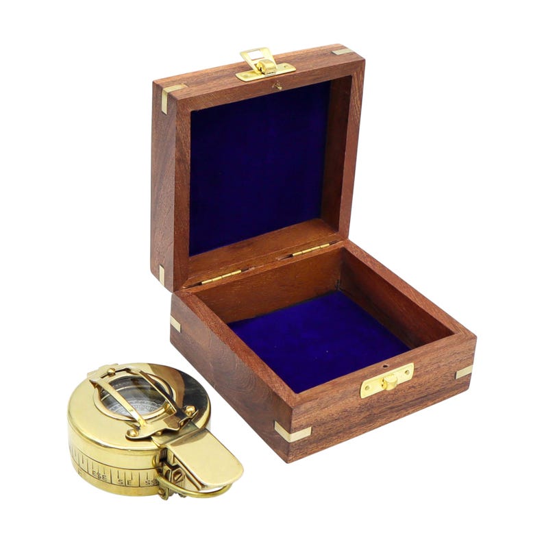 May include: A round brass compass with a hinged lid and a side latch, positioned next to an open square wooden box. The box features brass corner accents, a brass latch, and a deep blue velvet lining on the interior base and lid interior.