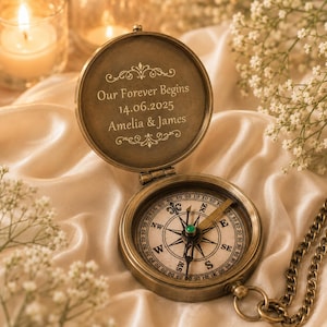 May include: A brass-toned pocket compass with an open lid featuring the engraved text Our Forever Begins 14.06.2025 Amelia & James. The compass face displays cardinal directions and a central needle, attached to a matching metal chain.