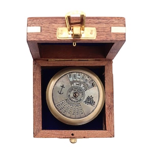 May include: A circular brass 100-year calendar with an anchor and ship engraving, housed in a square wooden box with a gold-toned metal latch. The calendar features numerical grids for years, months, and days, with text reading 2003-2099.