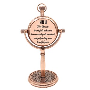 May include: A copper-toned metal desk clock with a circular frame mounted on a pedestal base. The clock face features tally marks and the text: Love like ours doesn't fade with time it becomes art shaped, weathered, and perfected by seven beautiful years.