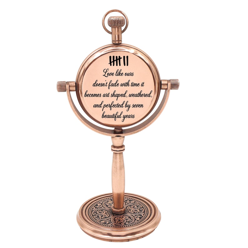 May include: A copper-toned metal desk clock with a circular frame mounted on a pedestal base. The clock face features tally marks and the text: Love like ours doesn't fade with time it becomes art shaped, weathered, and perfected by seven beautiful years.