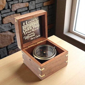 Può includere: Una scatola di legno aperta con una bussola all'interno. La bussola ha un quadrante nero e dettagli dorati. L'interno del coperchio recita: "Non sottovalutare mai la differenza che hai fatto e le vite che hai toccato. Buona pensione Harry Clarke."