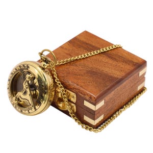 May include: A round brass sundial compass with a hinged gnomon and Roman numerals, attached to a gold-toned metal chain. The compass rests beside a square, polished wooden box featuring brass corner accents and a central brass latch closure.