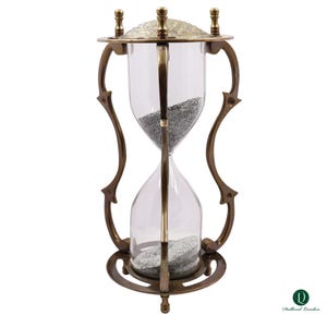 Puede incluir: Un reloj de arena de latón antiguo con cámaras de vidrio transparente. El reloj de arena está lleno de arena gris y tiene un marco de latón ornamentado con curvas y pináculos decorativos. La base tiene tres pequeños pies.