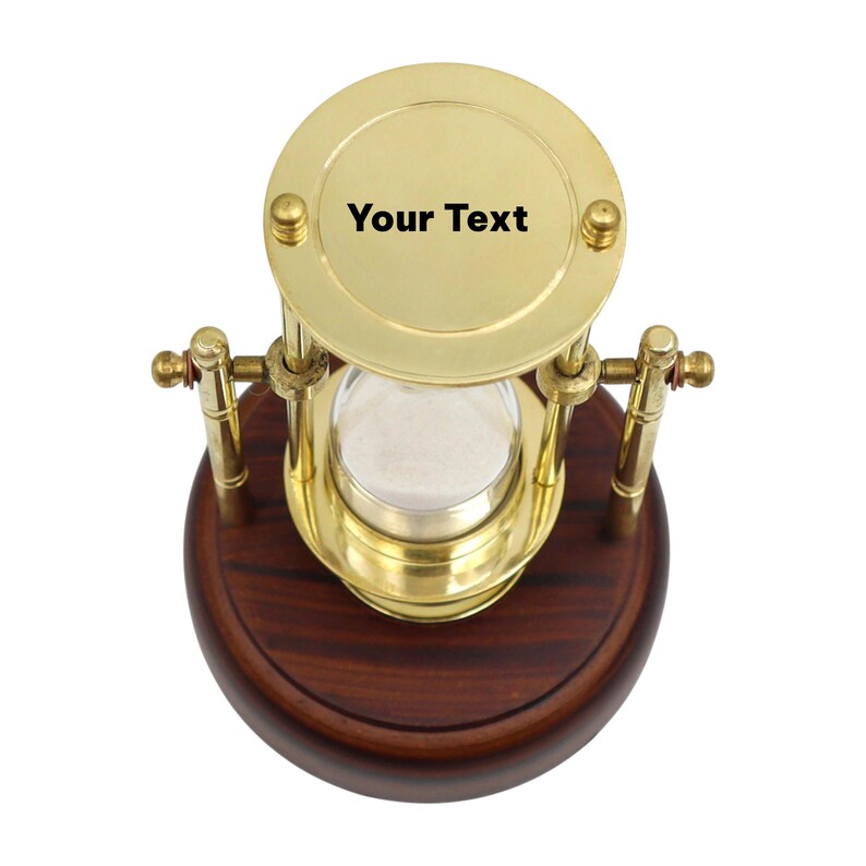 K&ouml;nnte beinhalten: Eine Messing-Sanduhr mit dem Text "Your Text" oben, auf einem dunklen Holzsockel. Die Sanduhr hat eine transparente Glaskammer, die mit wei&szlig;em Sand gef&uuml;llt ist. Die Messingkomponenten haben eine polierte Oberfl&auml;che.