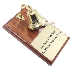 Puede incluir: Una campana de latón pulido montada sobre una placa de madera oscura rectangular con un tirador de cuerda blanca. Una placa de latón rectangular está fijada a la madera, con el texto grabado: Your Name & Date Here, Let This Bell Tell Your Story.
