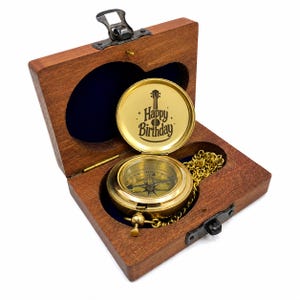 May include: A brass pocket compass with a gold-toned chain rests inside a dark brown wooden box. The interior lid of the compass features a black graphic of a guitar and the text Happy Birthday. The box has a metal latch and dark blue lining.