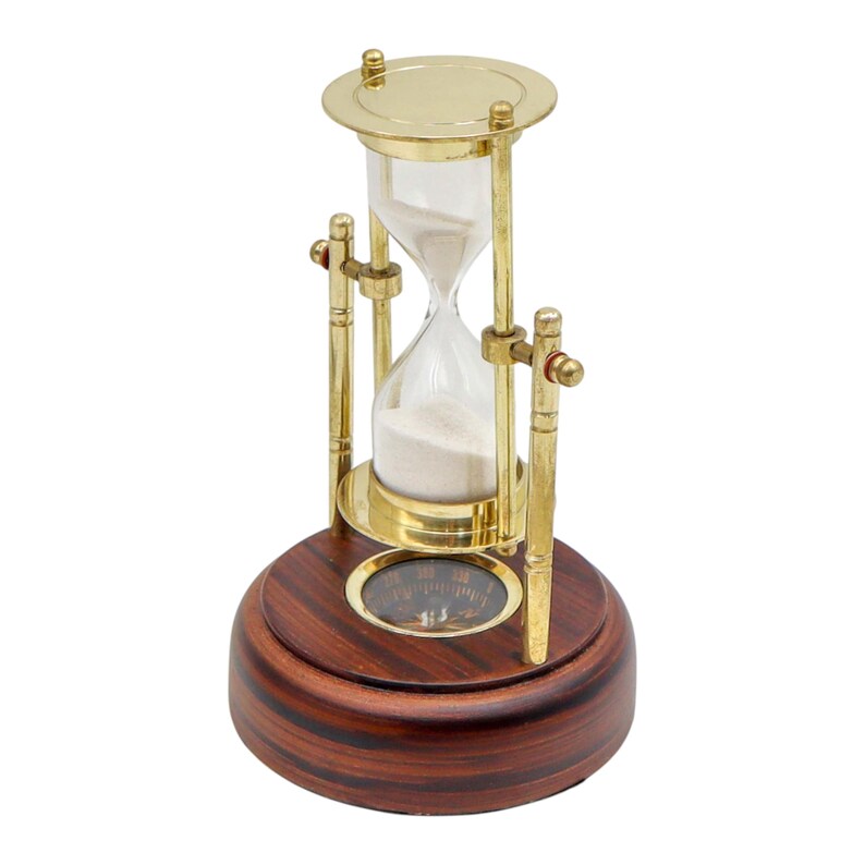 K&ouml;nnte beinhalten: Eine Sanduhr mit Messingrahmen und Holzsockel. Die Glas-Sanduhr enth&auml;lt wei&szlig;en Sand. Der Sockel hat einen Kompass. Der Messingrahmen tr&auml;gt die Glas-Sanduhr. Der Holzsockel ist dunkelbraun.