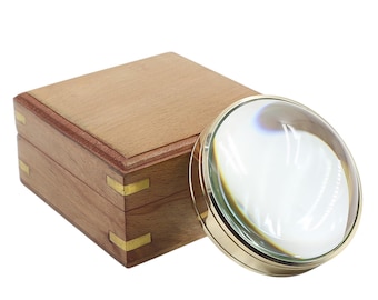 May include: A round glass paperweight magnifier with a polished gold-tone metal rim, positioned next to a square wooden box featuring brass corner accents and a smooth, light brown finish on the lid and sides of the container.