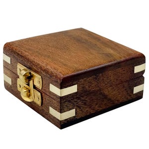 Może przedstawiać: Małe, kwadratowe drewniane pudełko z otwieraną pokrywą. Pudełko wykonane jest z ciemnobrązowego drewna z mosiężnymi narożnikami i mosiężnym zapięciem. Widoczne jest usłojenie drewna, a pudełko wydaje się być dobrze wykonane.