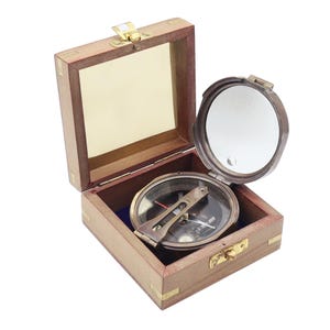 May include: A brass nautical compass with a hinged lid featuring an internal mirror, housed inside a square wooden box with brass corner accents and a metal latch. The compass sits on a blue interior lining within the open wooden container.