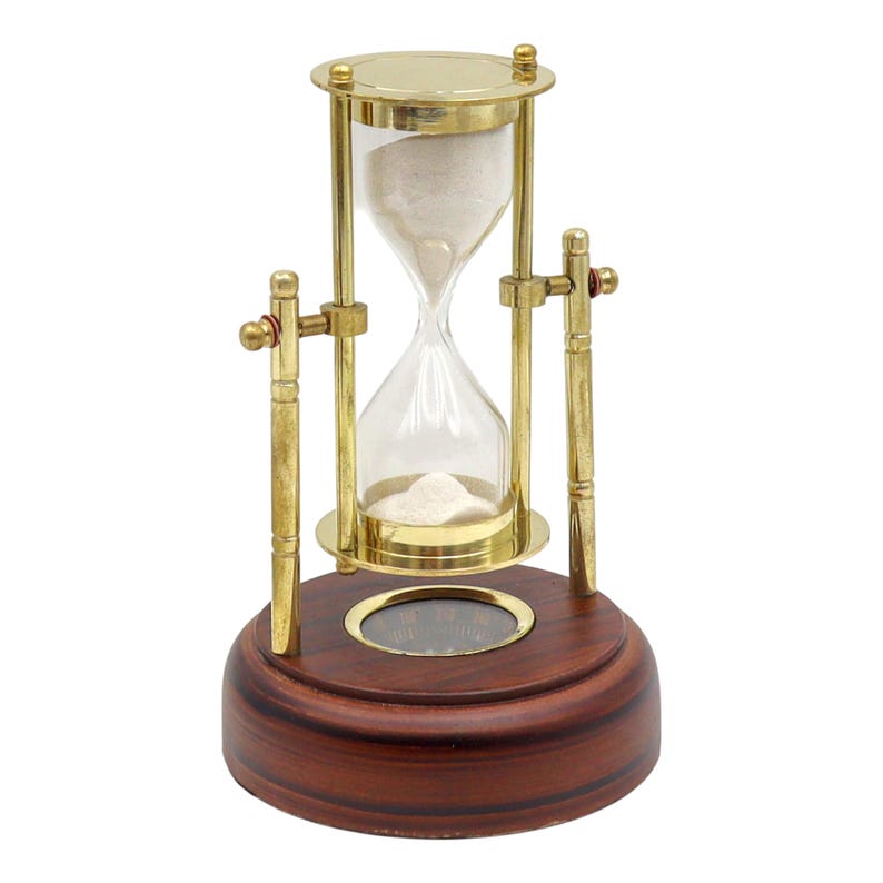 K&ouml;nnte beinhalten: Eine Sanduhr mit einem transparenten Glaskern, gef&uuml;llt mit wei&szlig;em Sand, in einem Messingrahmen. Die Sanduhr ist auf einem dunkelbraunen Holzsockel montiert. Ein klassisches Dekorationsst&uuml;ck.